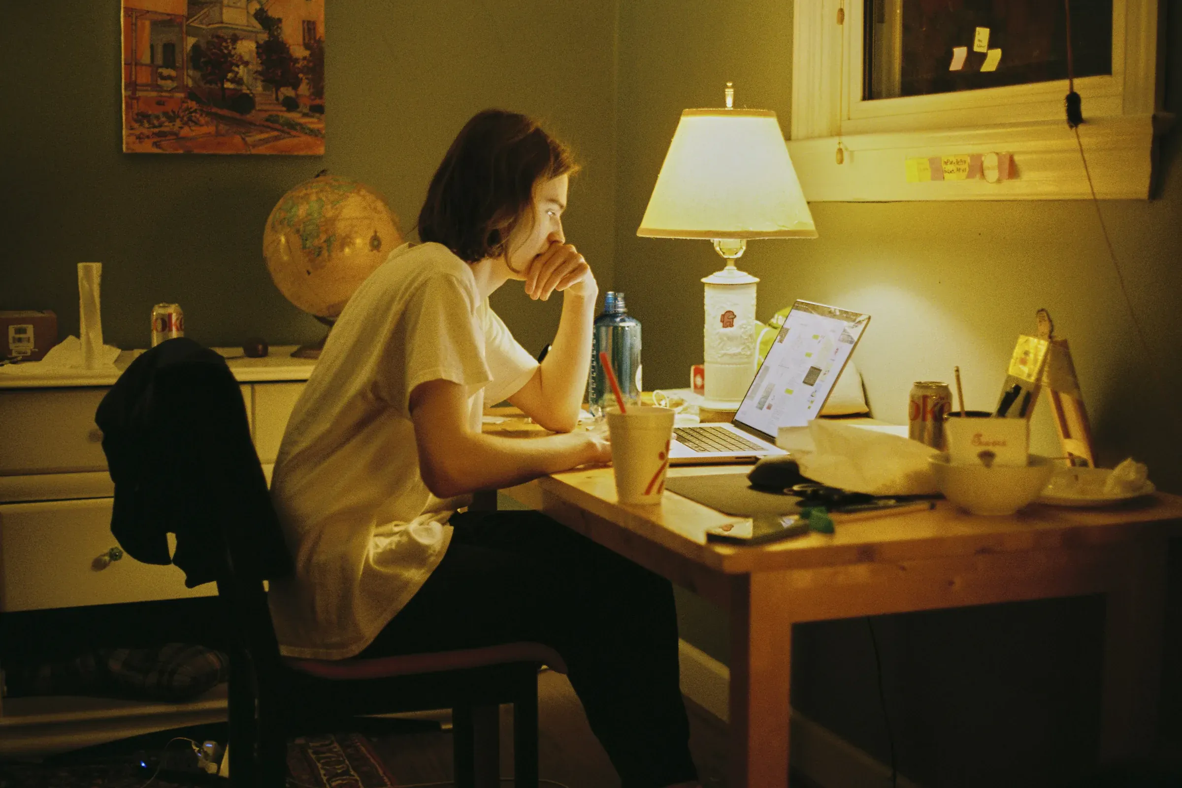A woman works at a desk by lamplight in a warm, cluttered study