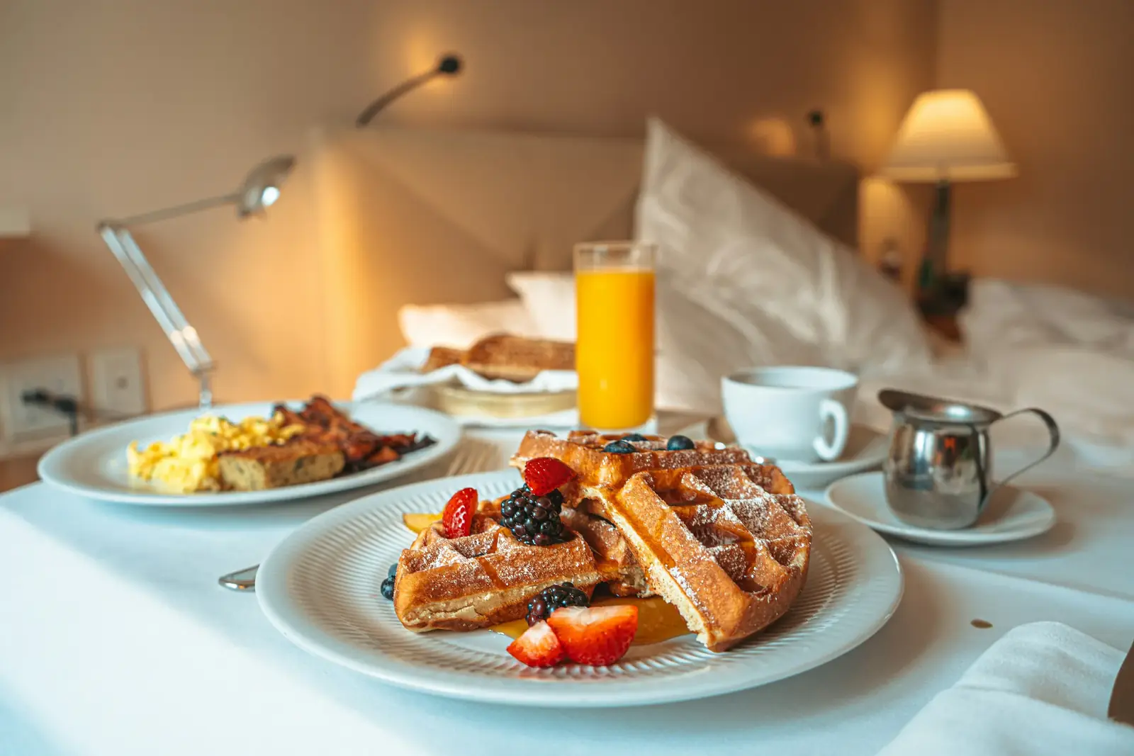 Breakfast in bed: waffles with berries, eggs and orange juice by a bedside lamp