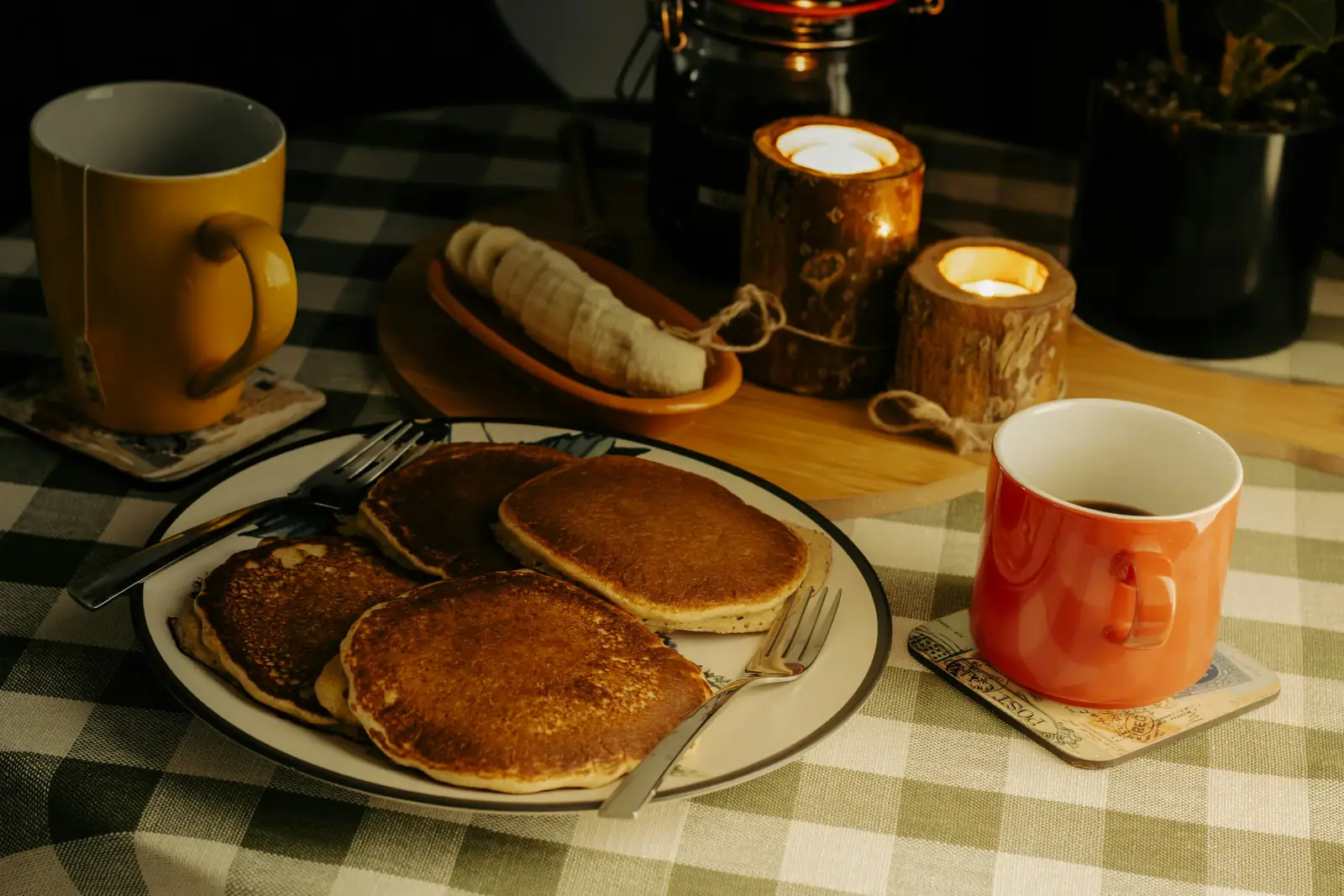 A plate of pancakes with mugs and candles on a checked tablecloth