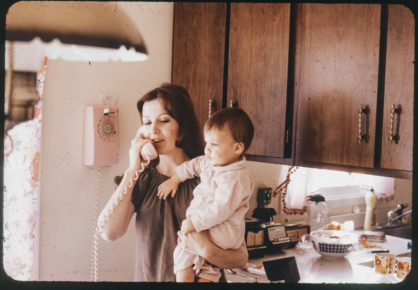 A woman holds her baby on her hip while talking on the phone in the kitchen