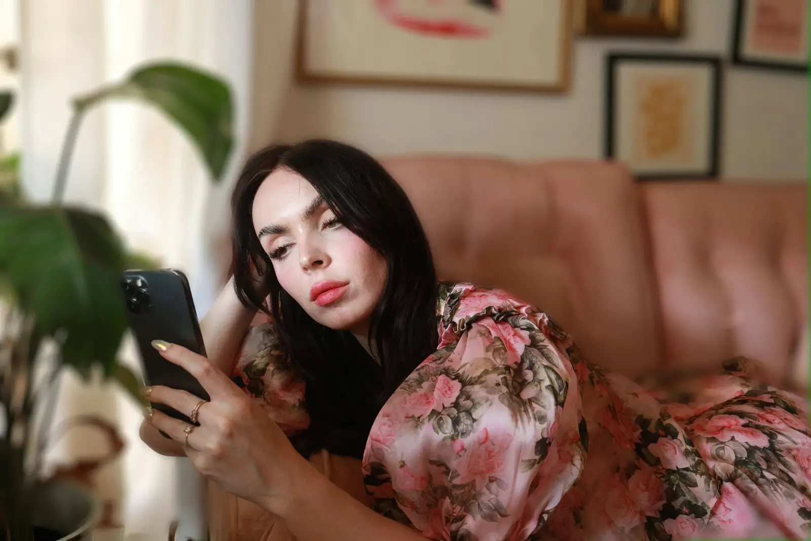 A woman on a pink sofa looks up from her phone with a guarded expression