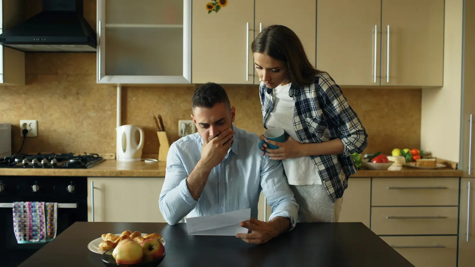 A man frowns over papers at a kitchen counter while his partner leans in beside him