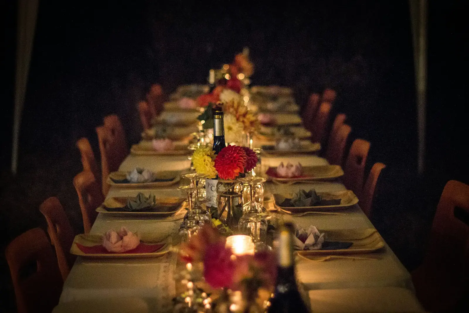 A long candlelit dinner table set for a party, with dahlias, wine glasses and folded napkins