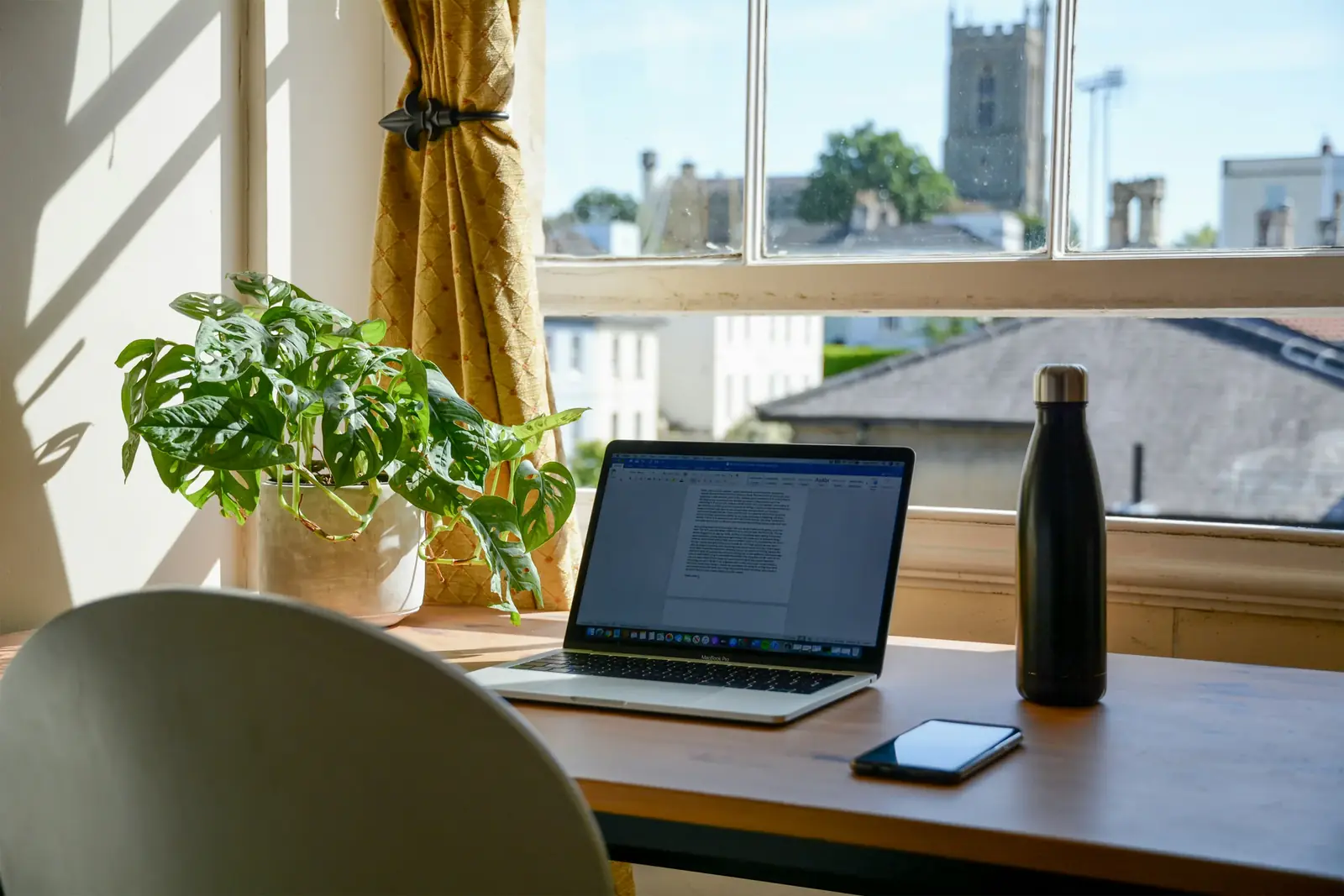 A laptop on a desk by a sunny sash window overlooking rooftops