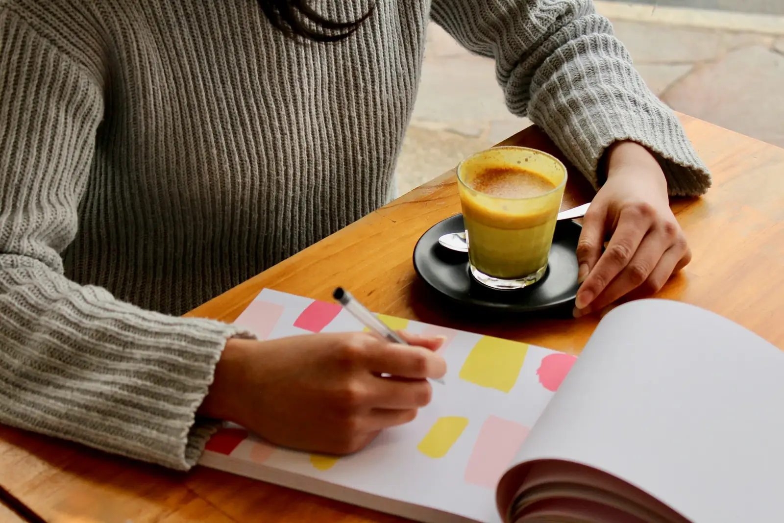 Hands writing a list in a notebook beside a cup of coffee on a wooden table