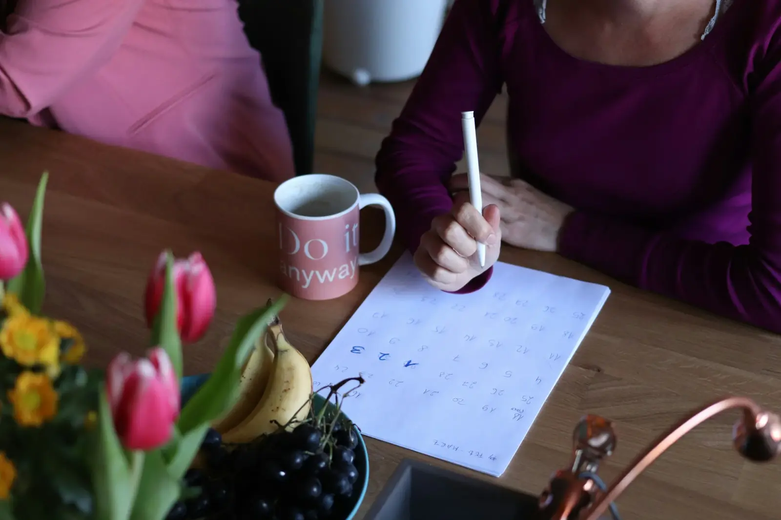 Hands writing in a planner at a kitchen table beside a mug and pink tulips