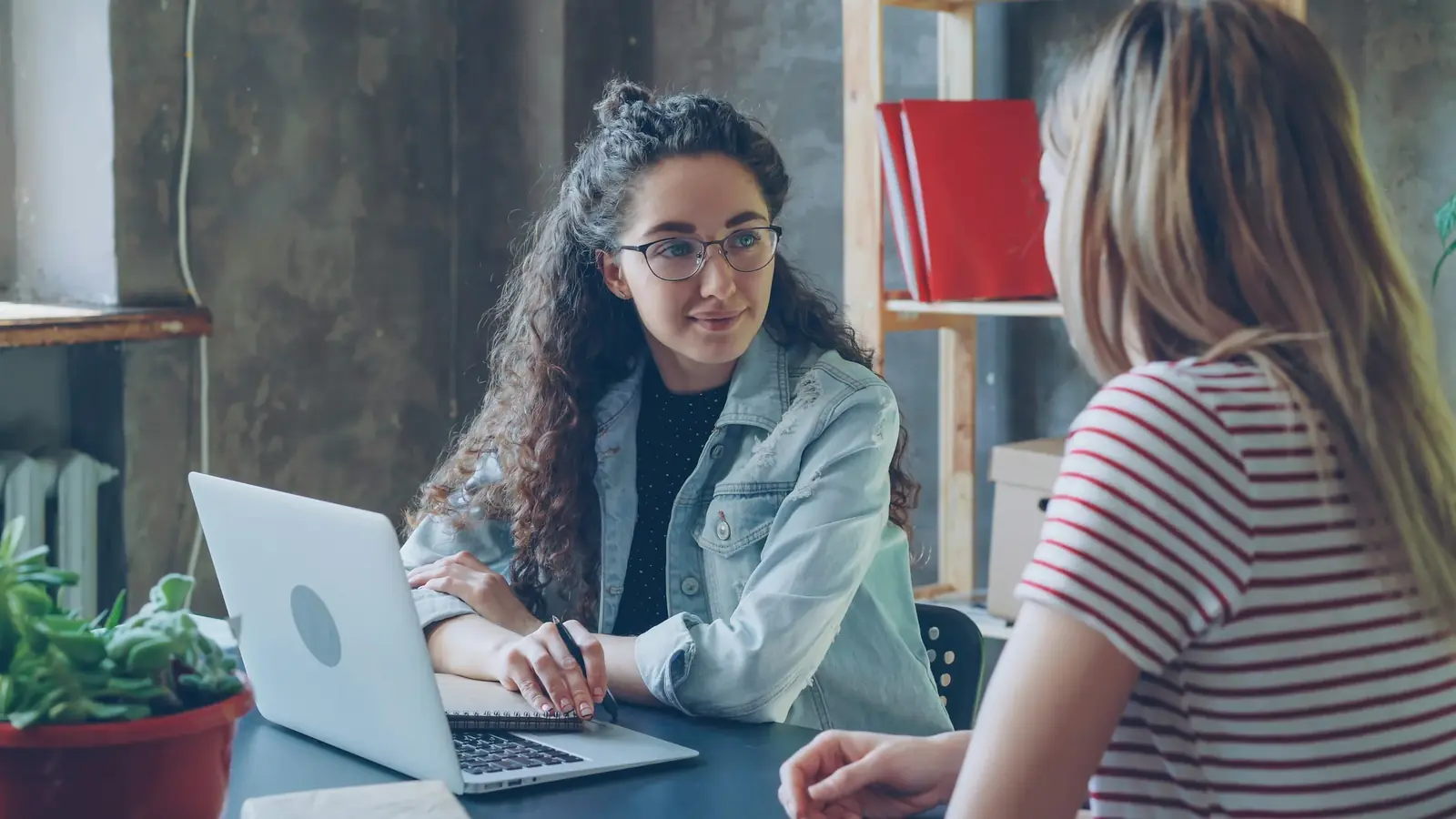 Two women talk at a café table, one with a laptop open