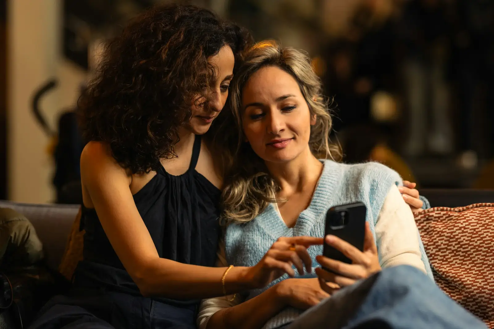 Two women sit close on a sofa in a warm, dim room, one scrolling her phone while the other looks on