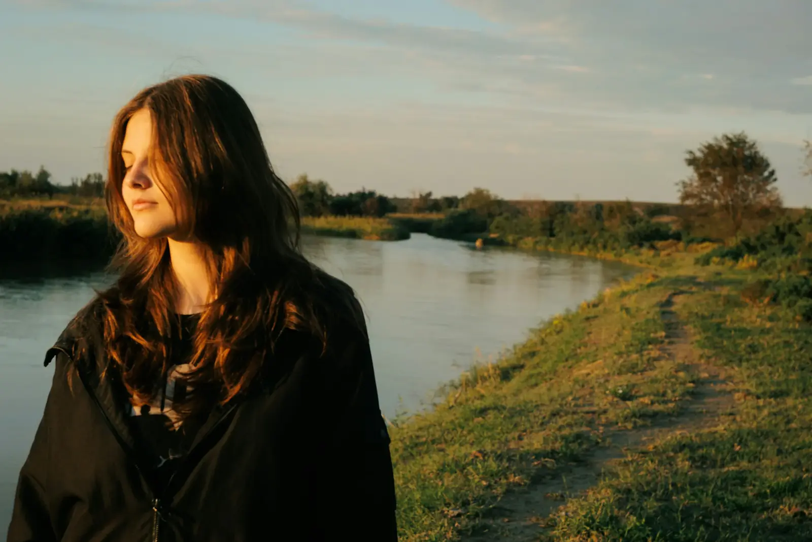 A woman with long dark hair stands by a river in late golden light