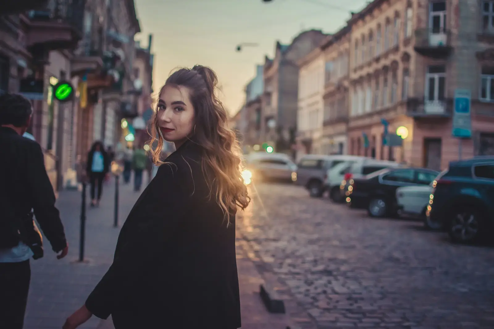 A woman in a dark coat glances back on a cobbled European street at dusk