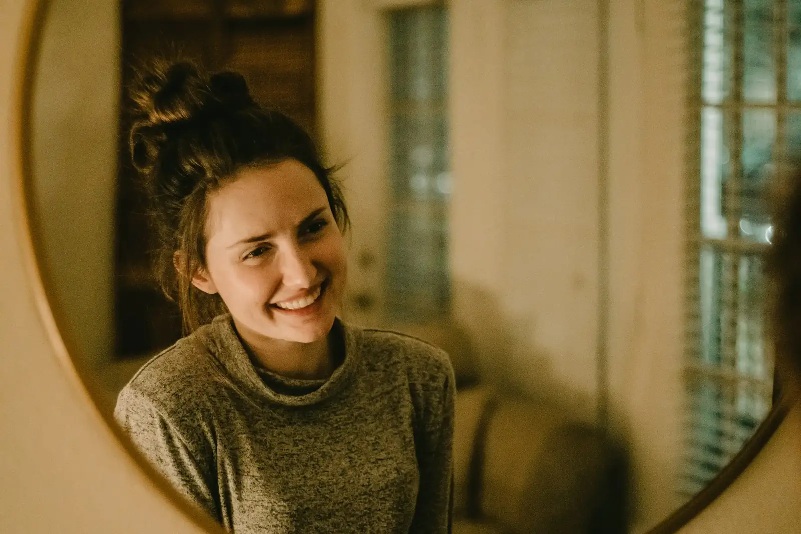 A woman smiles at her reflection in a round mirror in warm light