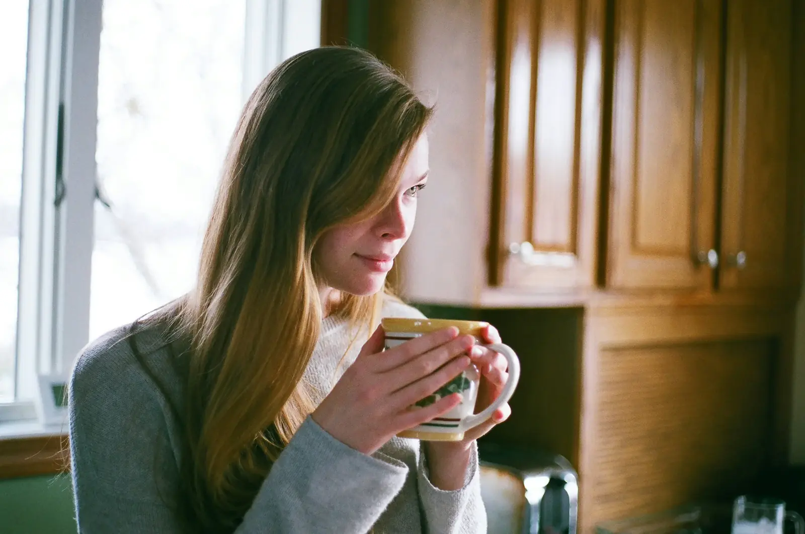 A woman in a grey sweater holds a warm mug by a sunny kitchen window