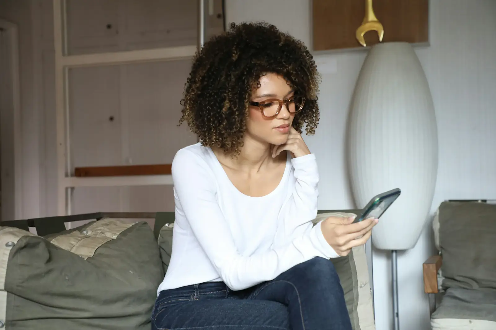 A woman with curly hair sits on a sofa looking down at her phone, chin in hand