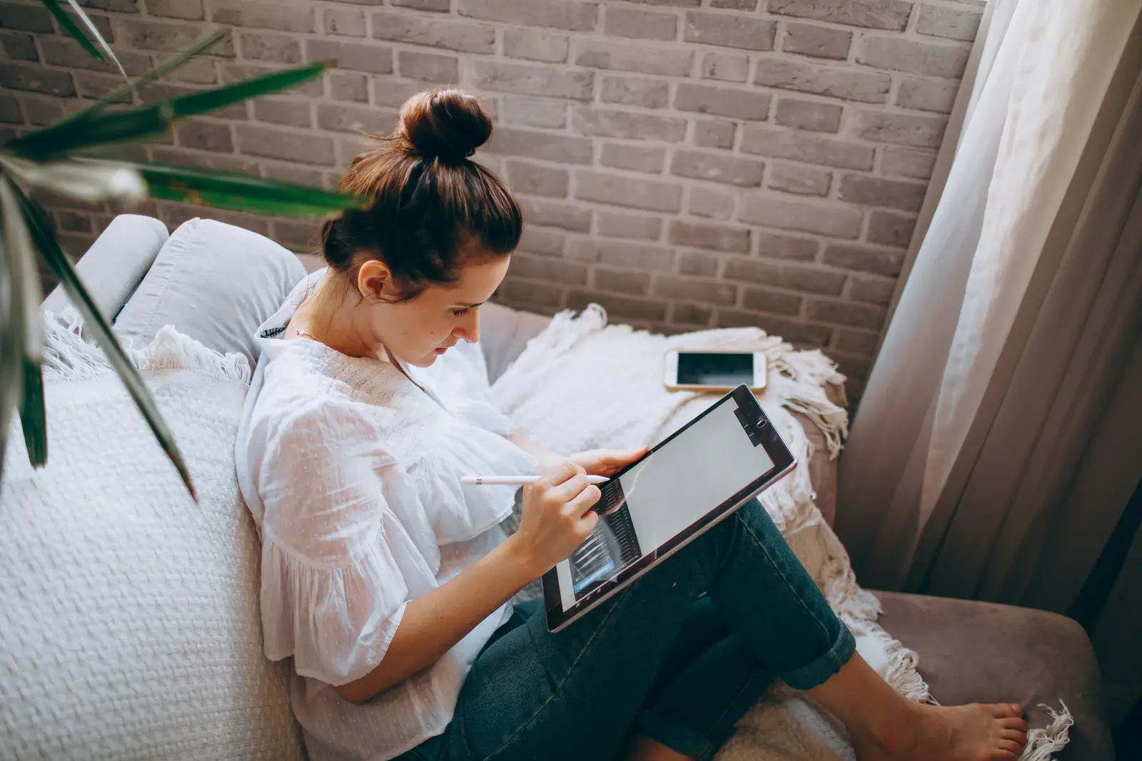 A woman with her hair in a bun works on a tablet on a white sofa by a brick wall