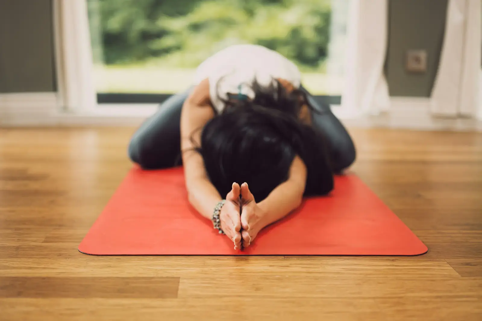 A woman rests in child's pose on a red yoga mat on a wooden floor