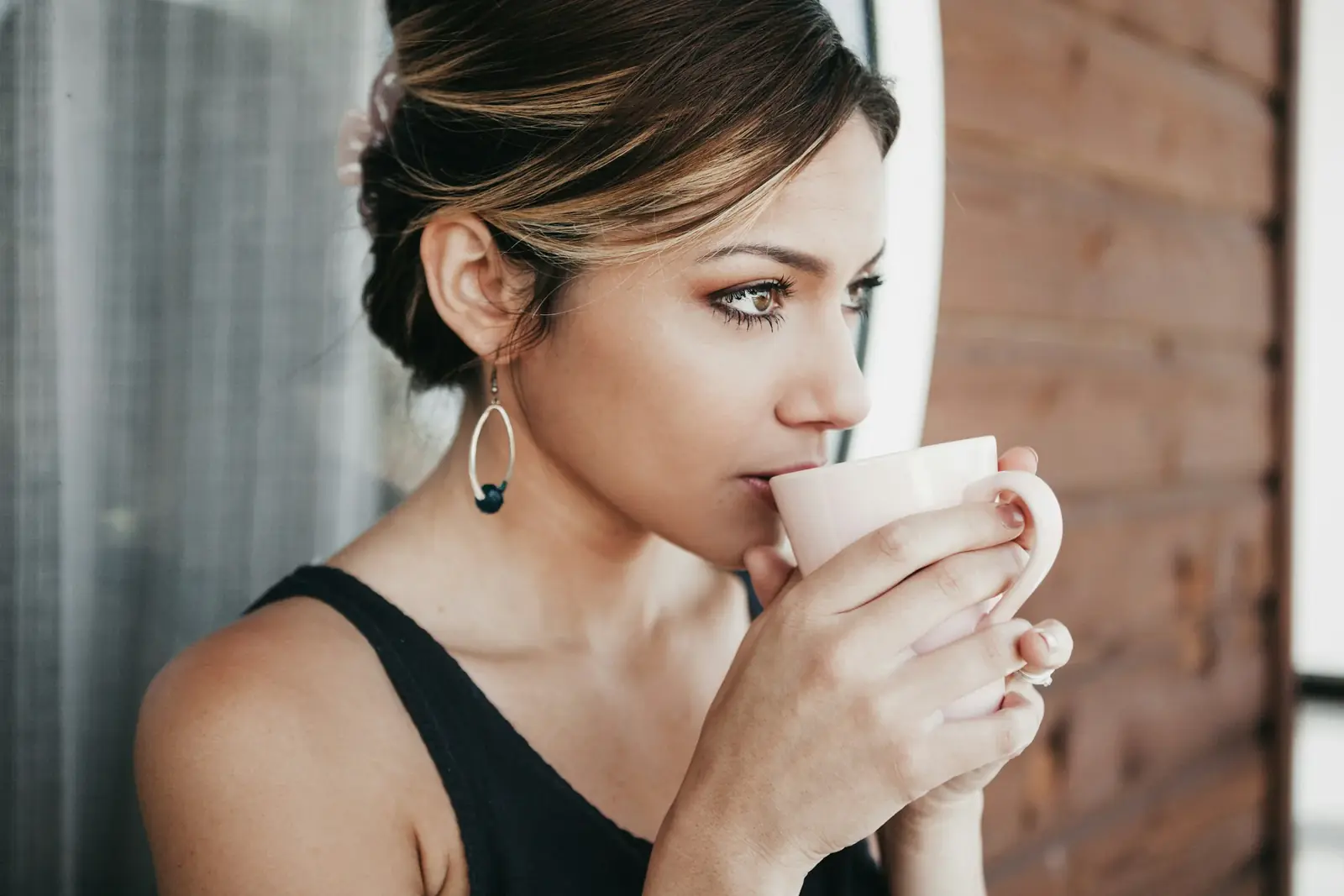 A woman sips from a mug with her eyes lowered by a brick wall