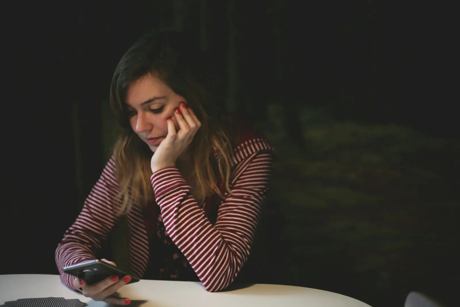 A woman leans on a table in a dark room, chin in hand, looking down at her phone