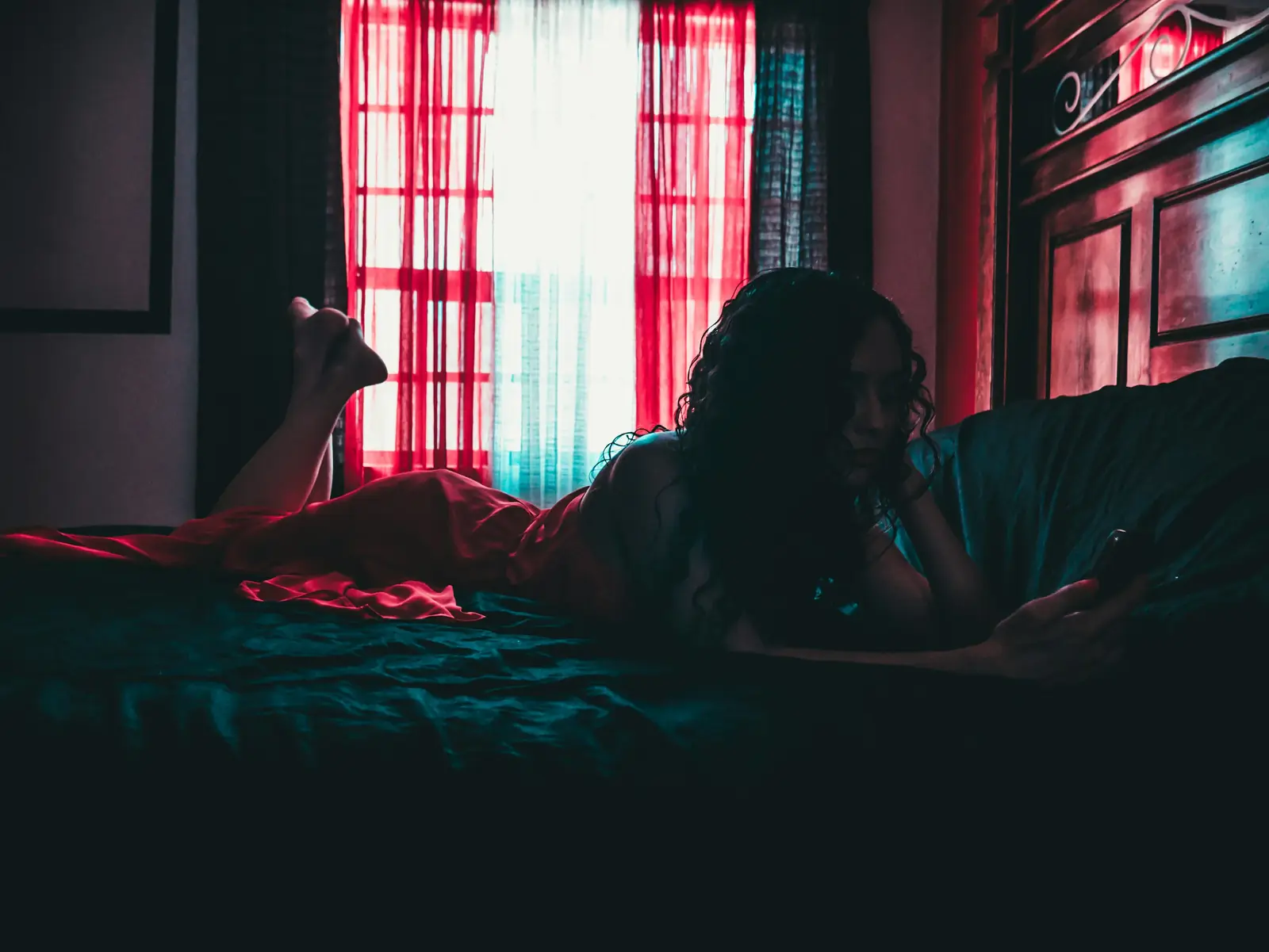 A woman lies on her bed in a dark room, lit by a window with red curtains, her phone in hand