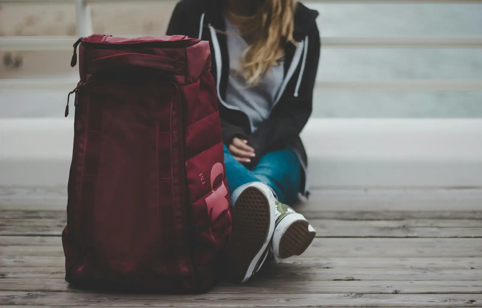 A person sits on a wooden floor beside a packed red backpack
