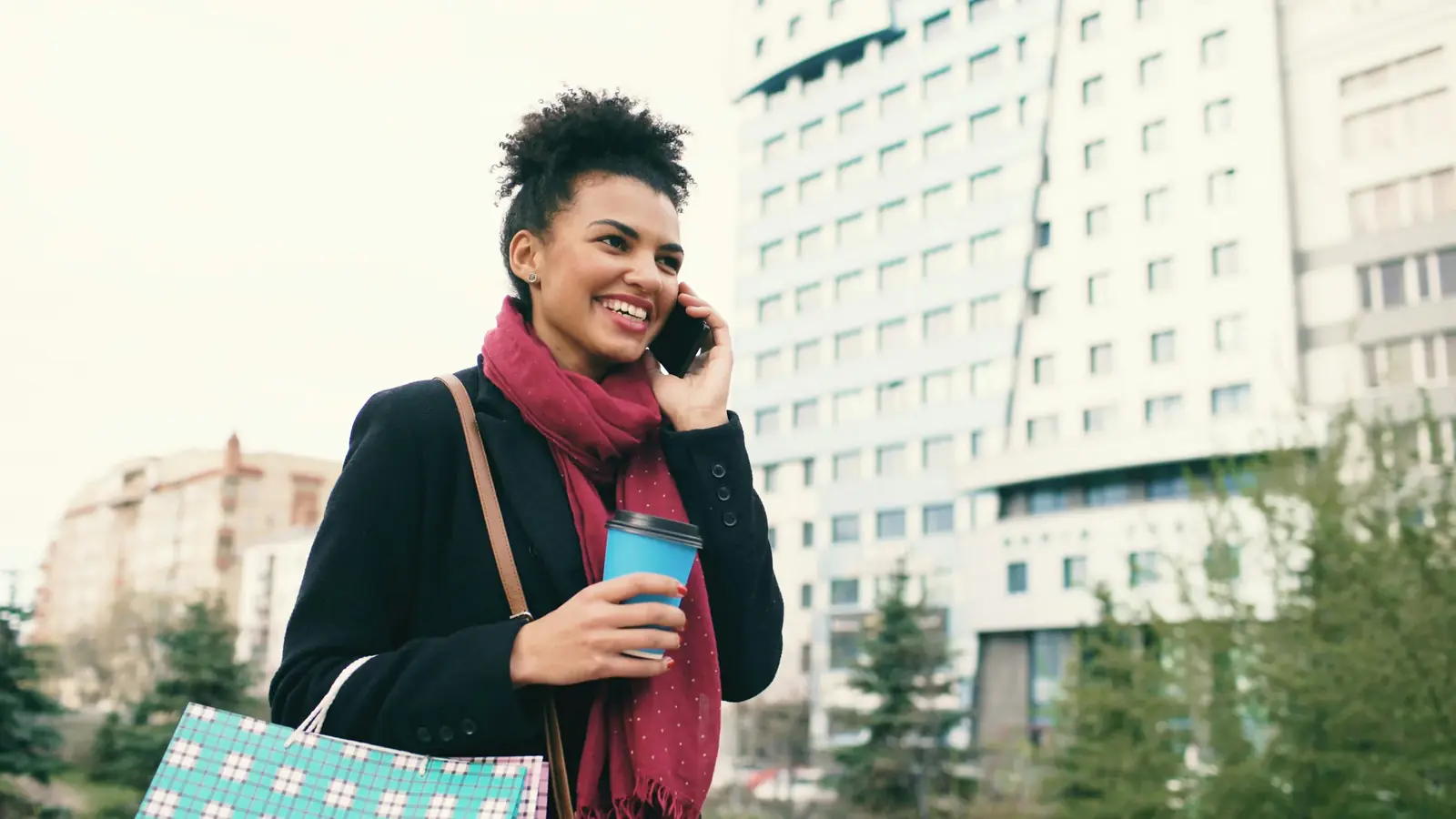 A woman in a red scarf talks on her phone, coffee in hand, on a city street