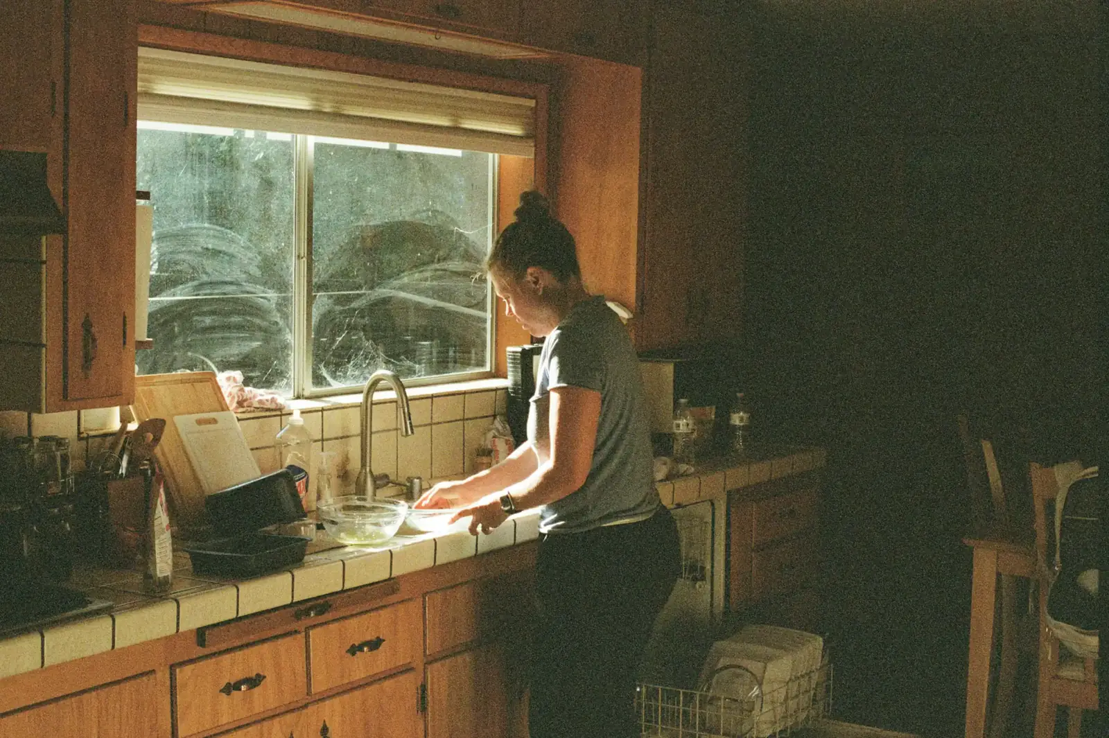 A woman washes up at a kitchen sink under a window in warm morning light
