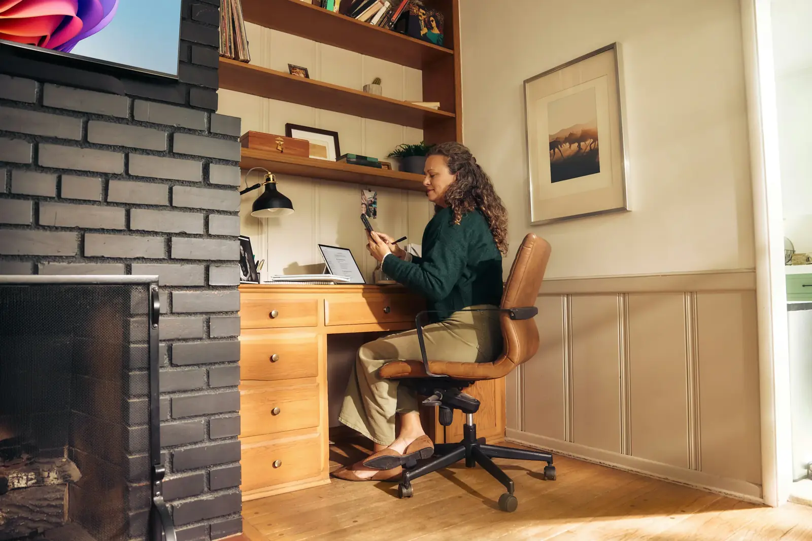 A woman works at a wooden desk in a sunlit home study with a brick wall