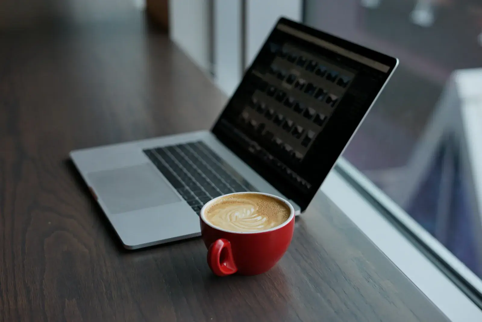 A laptop and a red coffee cup on a dark table by the window