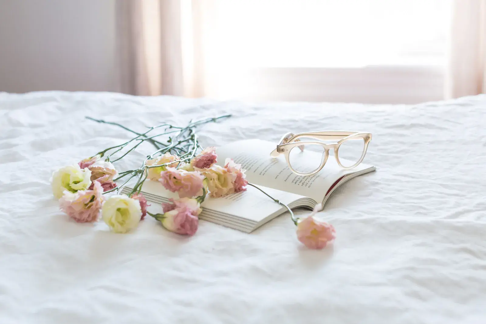Flowers, glasses and an open book on a white bed in morning light