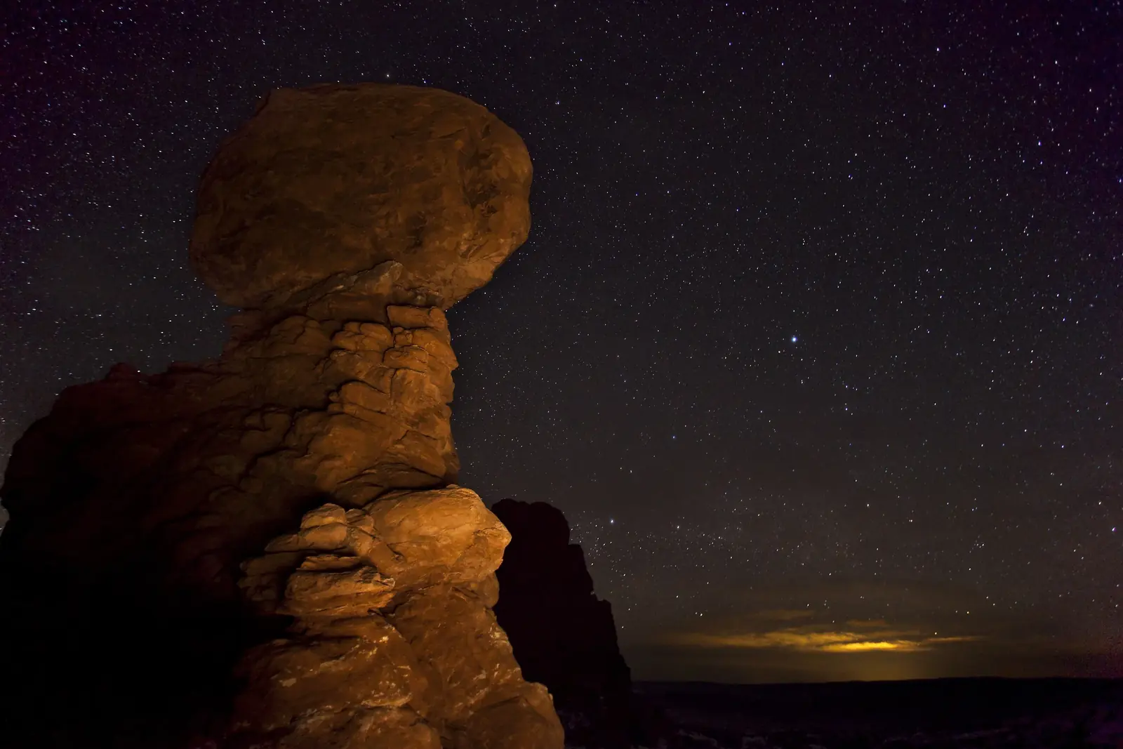 Balanced Rock lit warm against a starry predawn sky in Arches National Park