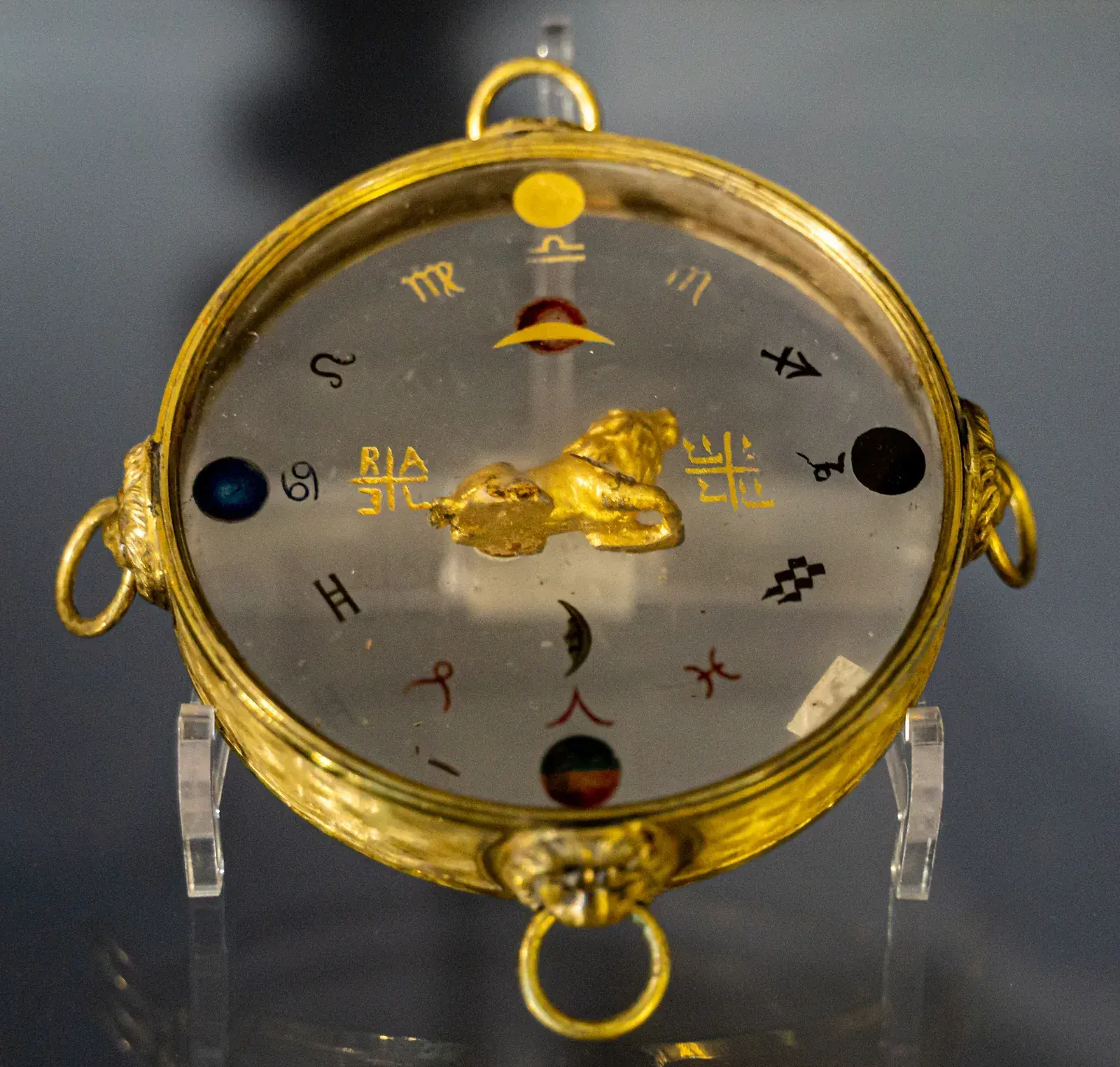 Round gilt amulet with a glass face painted with the twelve zodiac glyphs, coloured planet discs, a crescent and a small gold lion