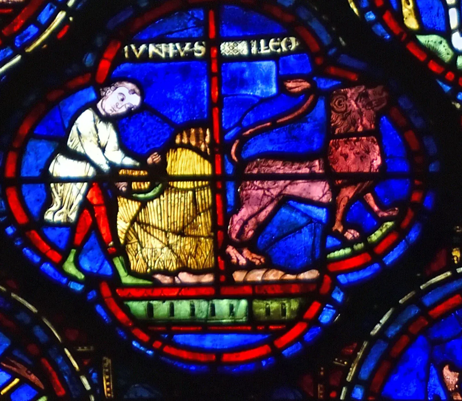 Stained-glass quatrefoil: a man in white working at a golden sheaf on the left, a red lion on the right, labelled IUNIUS and LEO