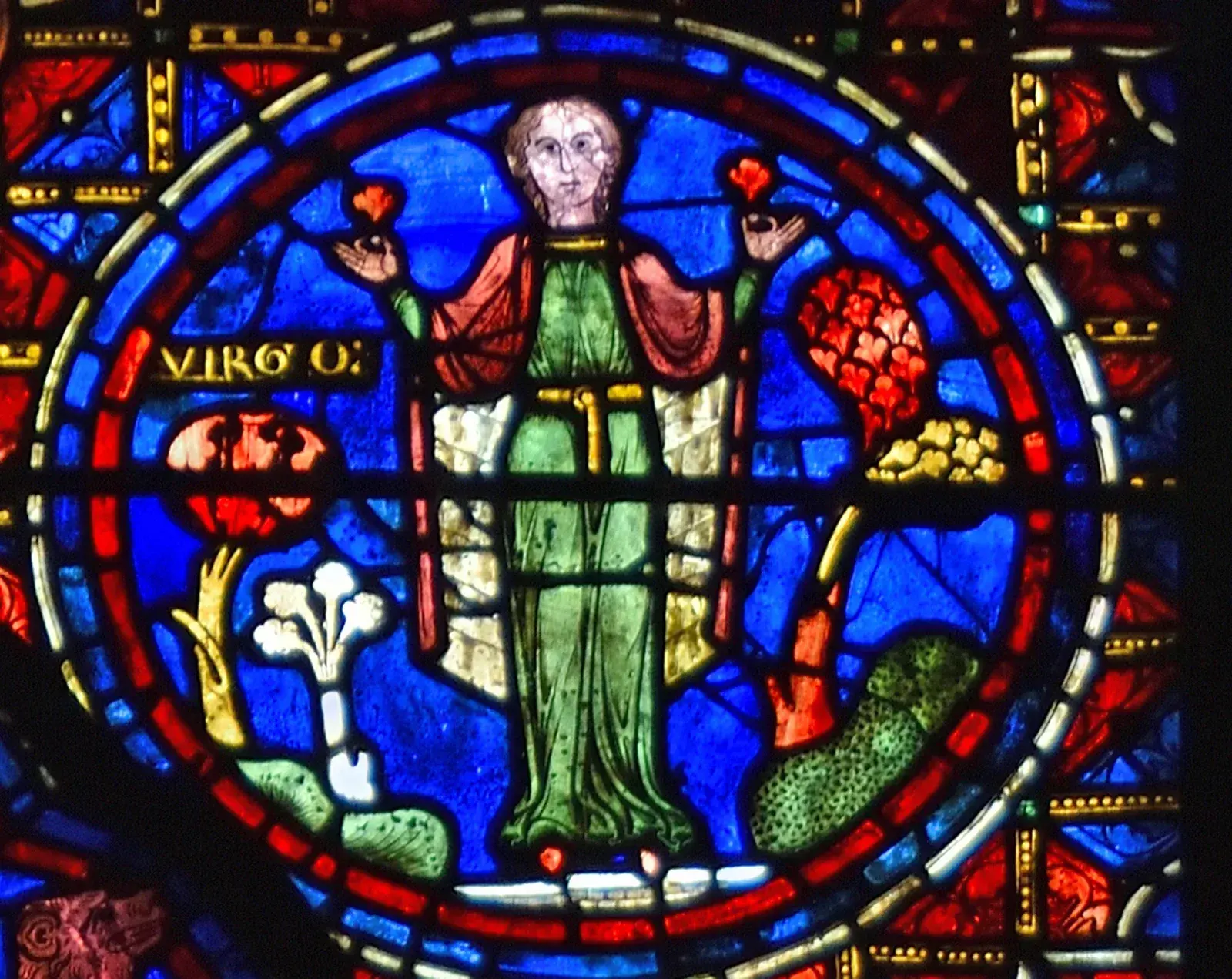 Stained-glass roundel of a robed woman holding flowers in both hands between fruiting plants, labelled VIRGO