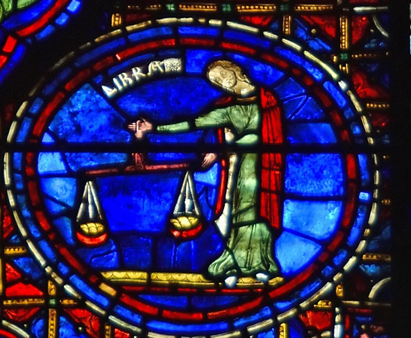 Stained-glass roundel of a robed figure holding a balance with two pans level, labelled LIBRA