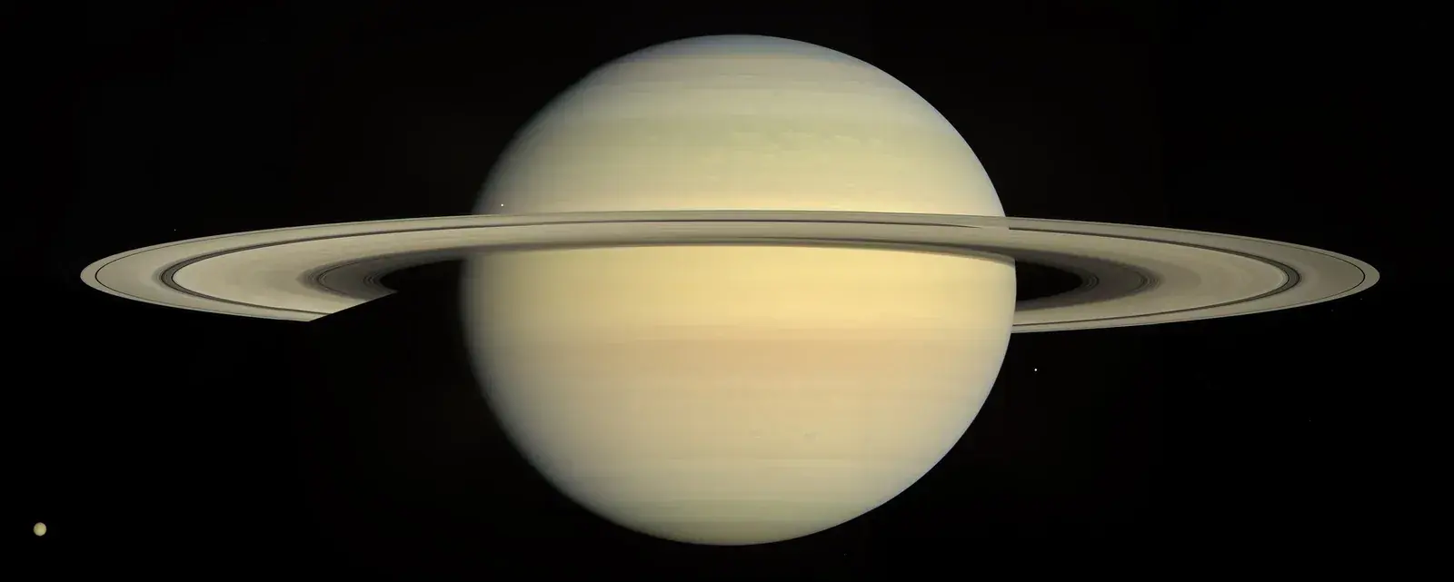Saturn in pale gold natural colour with its rings seen nearly edge-on and a small moon at lower left, on a black background