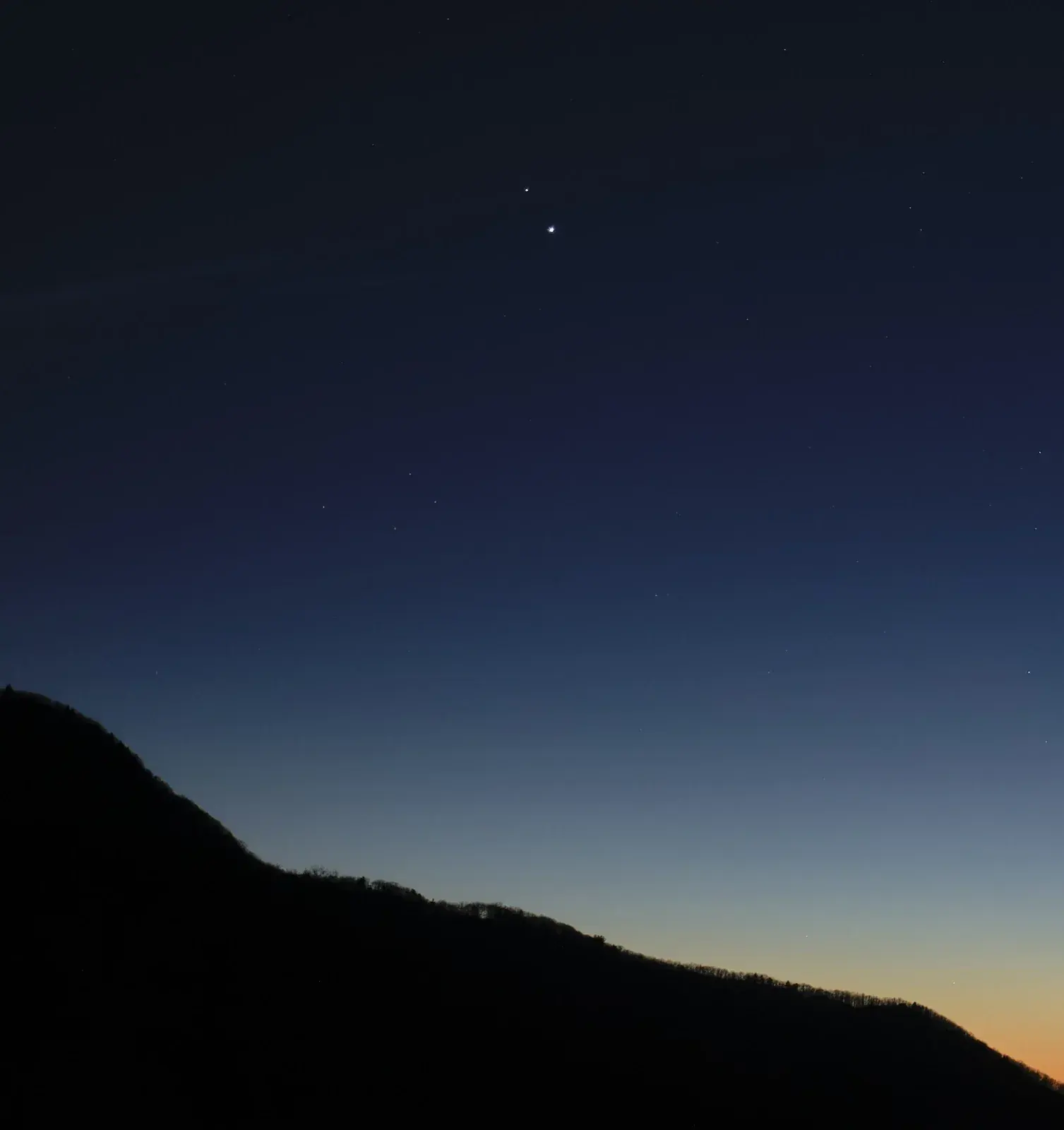 Twilight sky over a dark ridge with two bright points close together high on the left, Saturn above Jupiter