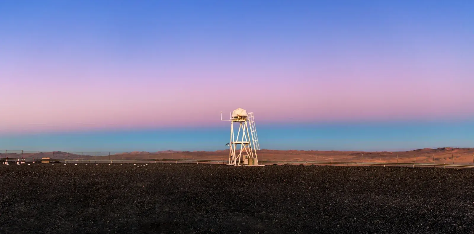 Twilight desert horizon with a dark blue band low in the sky under a pink band, and a white instrument tower in front
