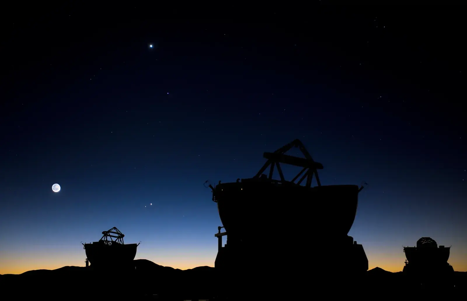 Dawn sky over silhouetted telescope domes with a crescent Moon on the left and four bright planets grouped in a short slanting line