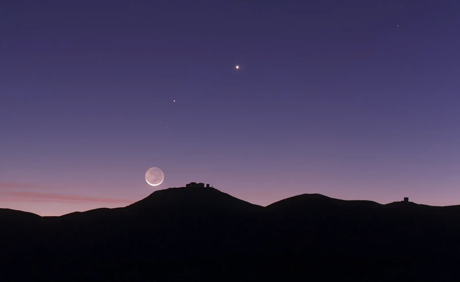 Thin crescent Moon with faint earthshine low over a dark mountain ridge at dusk, with two bright planets higher in the purple sky
