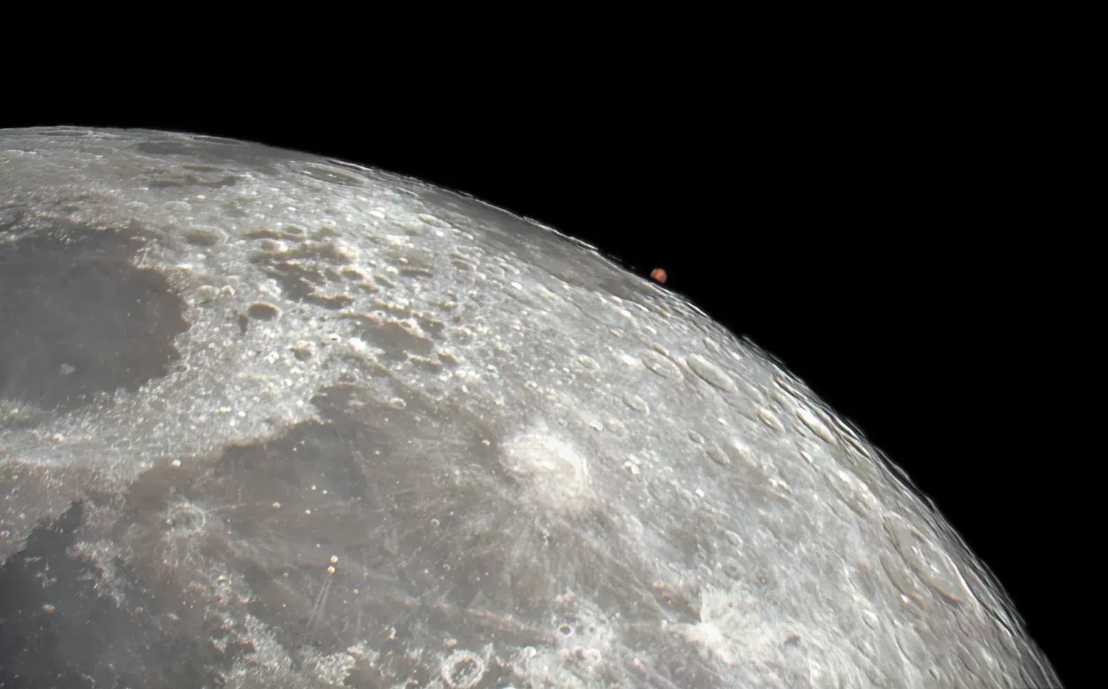 Telescope photo of the bright limb of the full Moon with small red Mars just emerging from behind its edge against black sky.