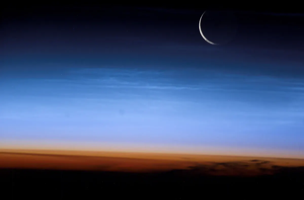A thin crescent Moon above the glowing blue layers of Earth's atmosphere, seen from orbit