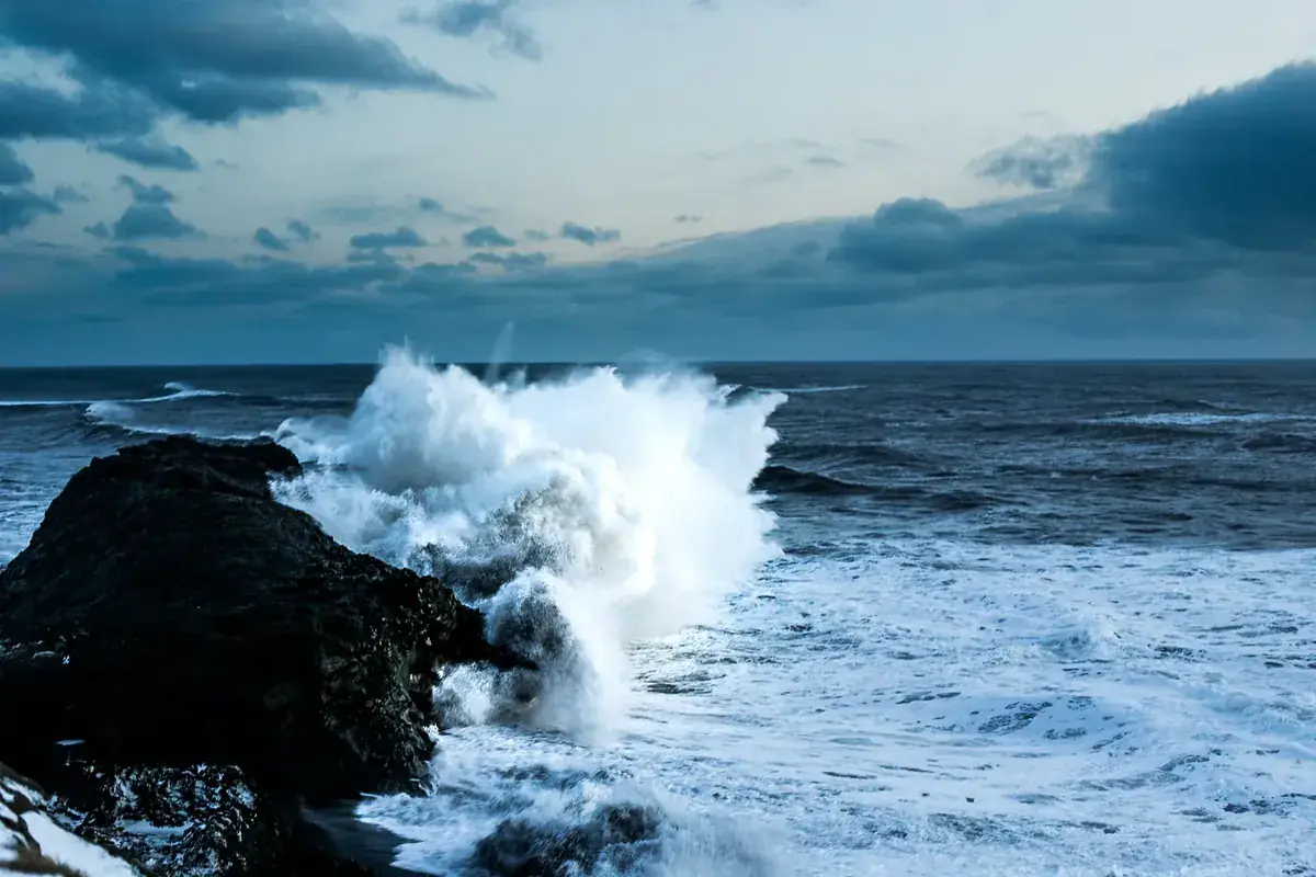 A wave breaks white against dark rocks under a stormy sea sky (Aries and Cancer compatibility)