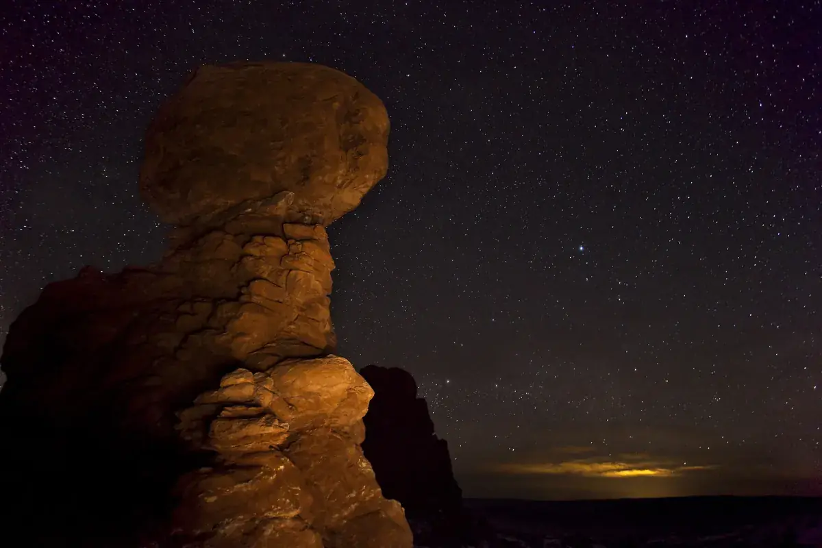 Balanced Rock lit warm against a starry predawn sky in Arches National Park